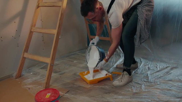 Young menders working hard, one of the repairers stands on the wooden ladder waiting for assistance, the other one pours out the white paint, mixing it. Workaholics, craftsmen. Slow motion, camera