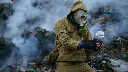 a man in a gas mask holds a sprout in the flask, against the background of fuming debris