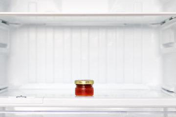 The delicates in a small glass in an empty refrgerator. A small bottle of jam in a shelf in the fridge. © milkovasa