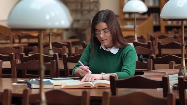Smart young woman in glasses taking notes while revising for exam, wearing smart white blouse and green sweater, indoor shot in old university library