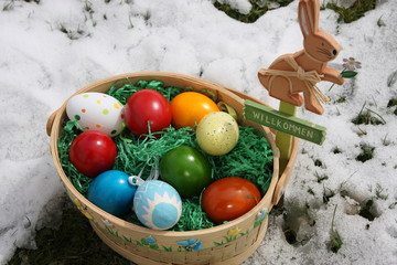 osterkorb mit bunten eiern und osterhase mit willkommensschild auf schneebedeckter wiese