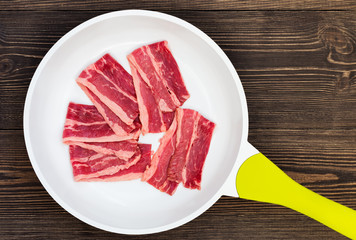 Bacon from marble beef, sliced, raw meat, lies on a white frying pan, with a yellow handle, on a dark background of natural wood
