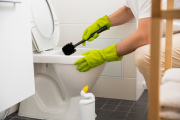 Man in rubber protective gloves cleaning and washing toilet with brush and bottle of cleaning agent in hands. White modern bathroom. Green liquid detergent dripping in toilet pan