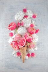 Flatlay waffle sweet ice cream cone with pink tulips and roses blossom flowers over white wood background, top view. Spring or summer mood concept.