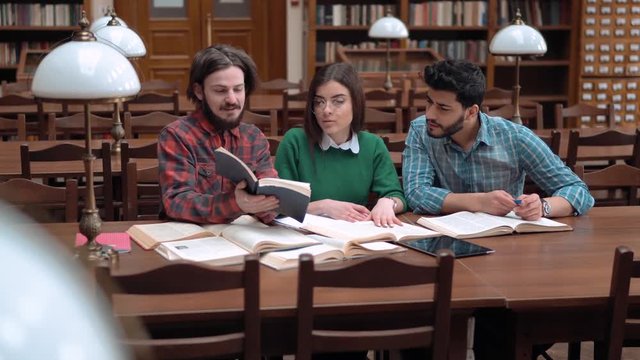 Three students working on home assignment in team, intelligent fair-haired boy pointing at information in book, beautiful brunette taking notes in notebook, handsome asian man giving opinion on the