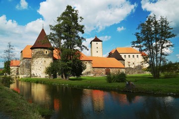 Fototapeta na wymiar Famous water castle Svihov at Czech republic, summer picture with moat, reflection in water, trees, green grass, blue sky, white clouds, palace, church, red roofs, rampart, beautiful scenery