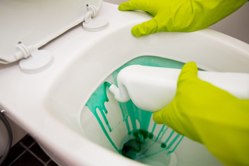 Man in rubber protective gloves cleaning and washing toilet with brush and bottle of cleaning agent in hands. White modern bathroom. Green liquid detergent dripping in toilet pan