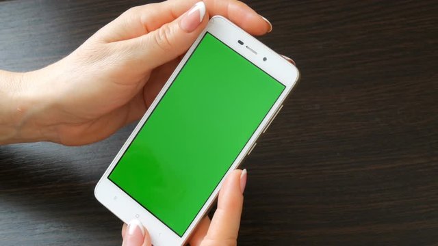 Female hands with beautiful French manicure take a white smartphone with Green Screen. Using Smartphone,Holding Smartphone with Green Screen on a stylish black wooden table