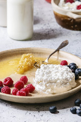 Ceramic plate of homemade cottage cheese served with blueberries, raspberries, bottle of milk and honeycombs over white marble texture table as background.