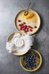 Fresh homemade cottage cheese in cheesecloth served in ceramic bowl with blueberries, raspberries and honeycombs over grey texture background. Top view, space