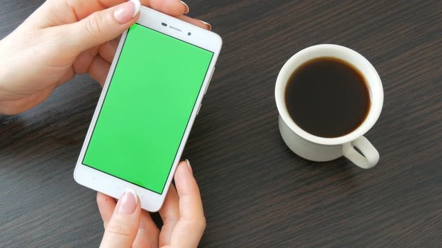Female hands with beautiful French manicure take a white smartphone with Green Screen near white cup of coffee. Using Smartphone,Holding Smartphone with Green Screen