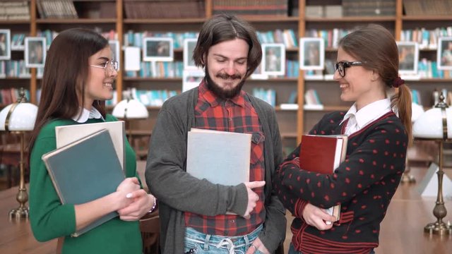 Happy students having lovely talk in library, two young women and bearded man chatting in front of bookshelves, holding books and study materials