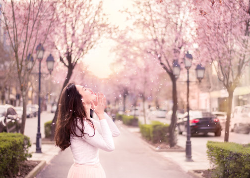 Young woman enjoying cherry tree petals falling to her face, happy emotion pink pastel colors, sunflare in background