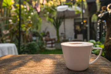 Cappuccino coffee in white cup on wooden table in garden with morning sun