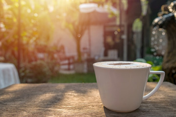 Cappuccino coffee in white cup on wooden table in garden with morning sun
