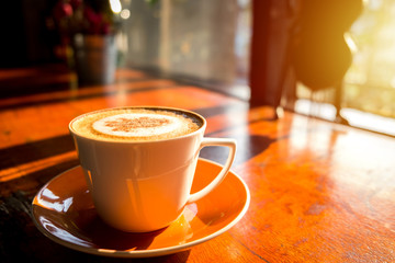 Cappuccino coffee in the white cup on wooden table with warm morning sun.