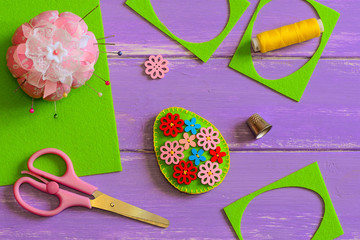 Simple felt Easter egg decoration idea. Hodemade felt Easter egg with colored wooden flower buttons. Felt scrap, scissors, thimble, thread, pincushion on the table. Easter projects. Easter crafts idea