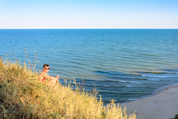 A young man in a red shirt sits on a hill in the background of the sea