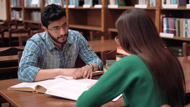 Two brilliant students discussing the issue while doing homework, handsome bearded boy and long-haired woman sitting at brown library table, giving high five after the talk