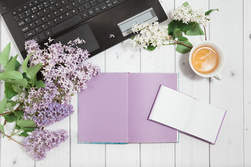 Beautiful branch of white and violet lilac flowers with opened note-book, pad and black opened laptop, lying on the white wooden background with cup of tea, mock up flat top view