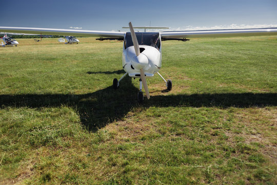 A small white light-engine aircraft with a propeller stands in the middle of a field with green grass on a summer clear sunny day.
