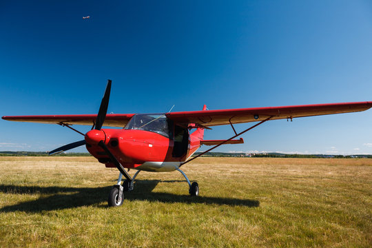 A small red light-engine aircraft with a propeller stands in the middle of a field with grass on a summer clear sunny day.
