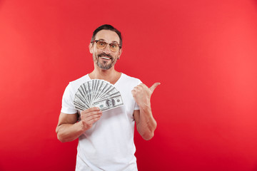 Handsome bearded man 30s in casual white t-shirt wearing glasses holding lots of money prize dollar currency and pointing finger aside on copyspace, isolated over red background