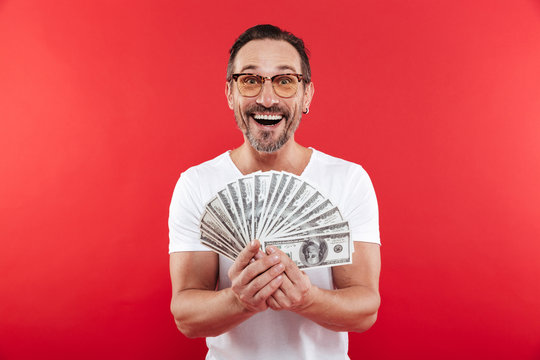 Photo of excited and rich man in casual white t-shirt smiling and demonstrating fan of money in dollar banknotes holding in hands, isolated over red wall