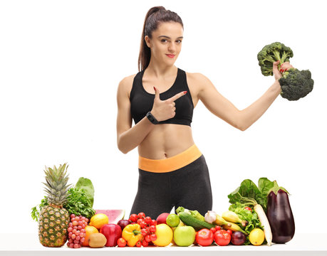 Fitness woman holding a broccoli dumbbell behind a table with fruit and vegetables and pointing