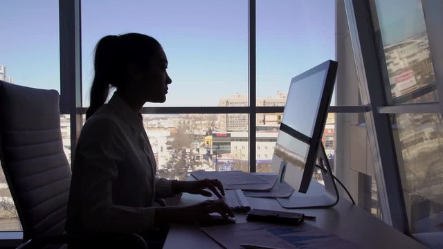 Female silhouette work on keyboard in front of monitor near window in office room during day. Concept: female professions, office time, landscape in window