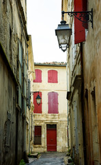 Fototapeta na wymiar Narrow the street in Arles (Provence, France). Weathered stucco walls, red wooden shutters and forging lanterns.