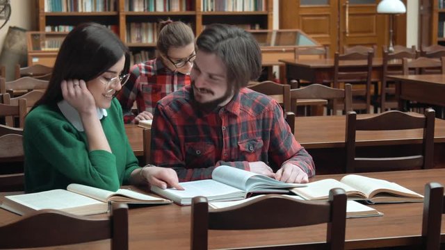 Two friends reading in big light library, smart bearded boy reading book, intelligent girl in glasses pointing at the text, having nice time after classes