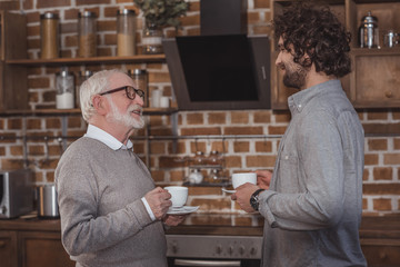 adult son and senior father standing with cups of coffee in kitchen