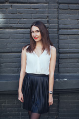 Young brunette woman with red lips wearing black skirt and white blouse smiling wide. Girl, black brick wall background, copy space, closeup.vertical