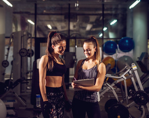 Fototapeta na wymiar Portrait view of young smiling attractive healthy fitness sporty active slim girl consulting with her pretty female personal trainer while holding clipboard and pencil in the gym.