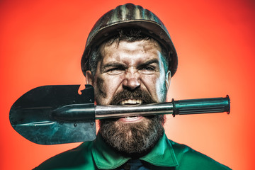 Mining and construction concept - man with angry face in protective helmet holds shovel in teeth....