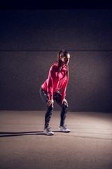 Portrait vertical view of young athletic slim fashion active girl crouching while having a break on the street in front of the grey wall at night.