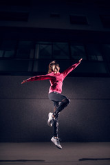 Portrait low vertical view of young athletic slim fashion active girl jumping with open arms on the street in front of the grey building at night.