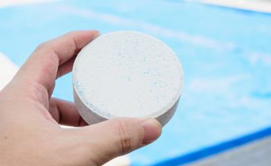 Hand holding a white rounded chlorine tablet. Unfocused swimming pool on the background.