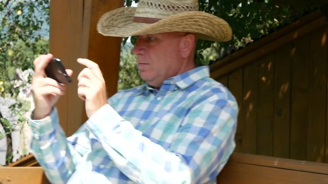 Farmer Sitting Outside in a Wooden Swinger Use Mobile Phone to Send Messages