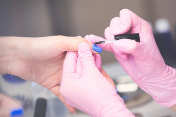 Closeup of manicurist in pink rubber gloves applying blue nail polish on female nails. Professional manicure and nail care procedure in beauty salon