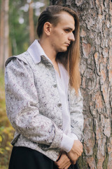 Sexy long-haired man with a pipe in his hands, in the woods. In a beautiful, ancient jacket with patterns and a white shirt. Fantasy.