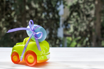 Easter light green car with a blue egg tied with a purple ribbon rides on the table against the background of greenery