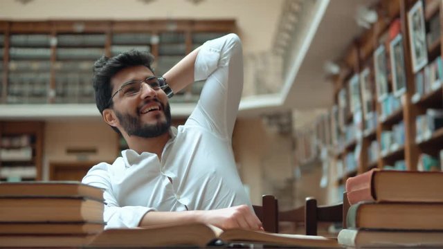 Smart student succeeds as reading a book, attractive young man in glasses understands the idea while doing homework, indoor shot in spacious rich library