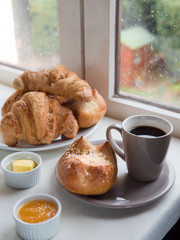 a brioche, French bun , croissants, with butter, jam and a cup of coffee in front of a window on a rainy day
