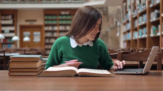 Bright female student studying in huge library, prettty girl in green sweater and smart white blouse searching information on the internet, using white laptop while reading encyclopedia
