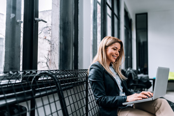 Portrait of a smiling blonde woman using laptop while sitting near the window.