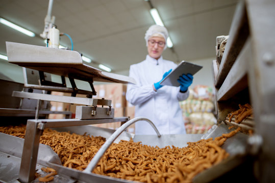 Close up view of a salt stick snacks being processed through a production line in food factory while being inspected by a female young worker with a tablet in a sterile cloth.