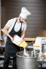 Handsome baker in uniform filling flour into the professional kneader for bread baking at the bakery