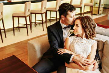 Bride and groom sitting at the bar on the couch. Cute married couple in cafe, groom kissing a bride. Pure tenderness
